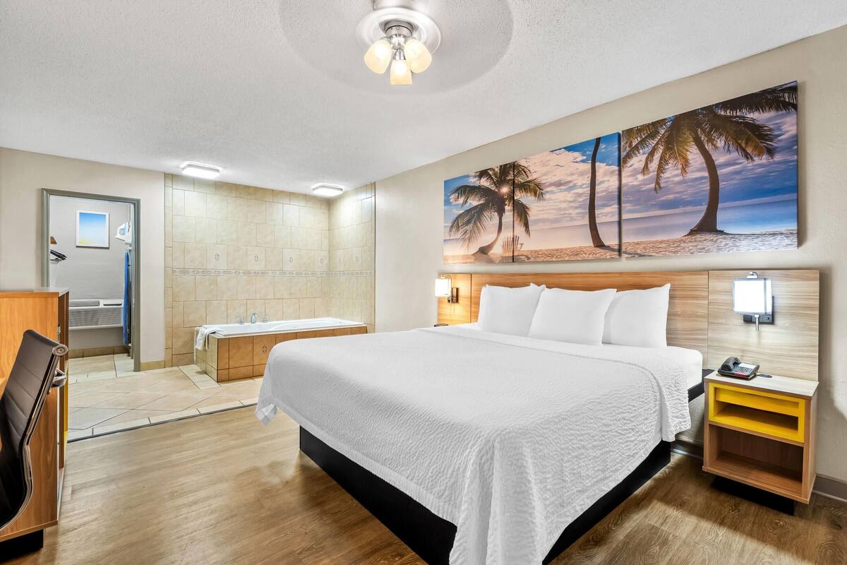 Ein Hotelzimmer mit einem großen Bett, einem Badezimmer mit Badewanne und einem Palmenbild an der Wand.