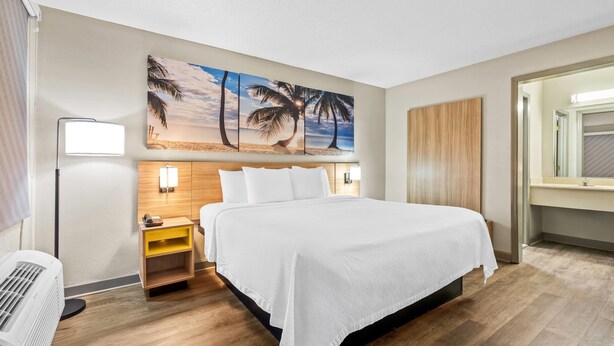 Una habitación de hotel con una cama grande, una mesita de noche, una lámpara de pared y un cuadro con palmeras.