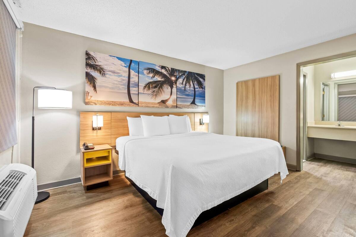 Ein Hotelzimmer mit einem großen Bett, einem Nachttisch, einer Wandleuchte und einem Palmen-Gemälde.