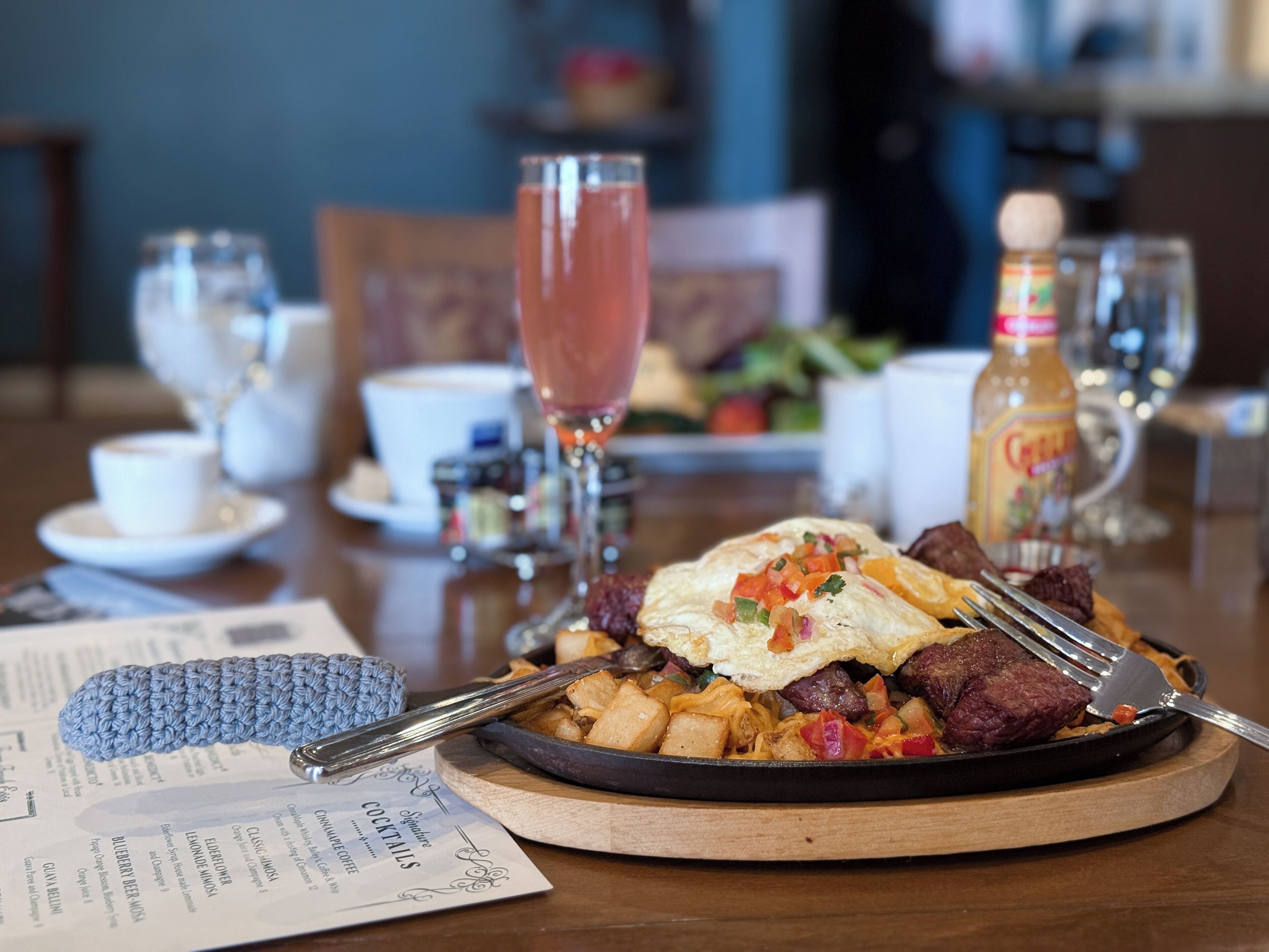 Un plato de desayuno con huevos, papas y carne, acompañado de una copa de cóctel y una botella de sirope.