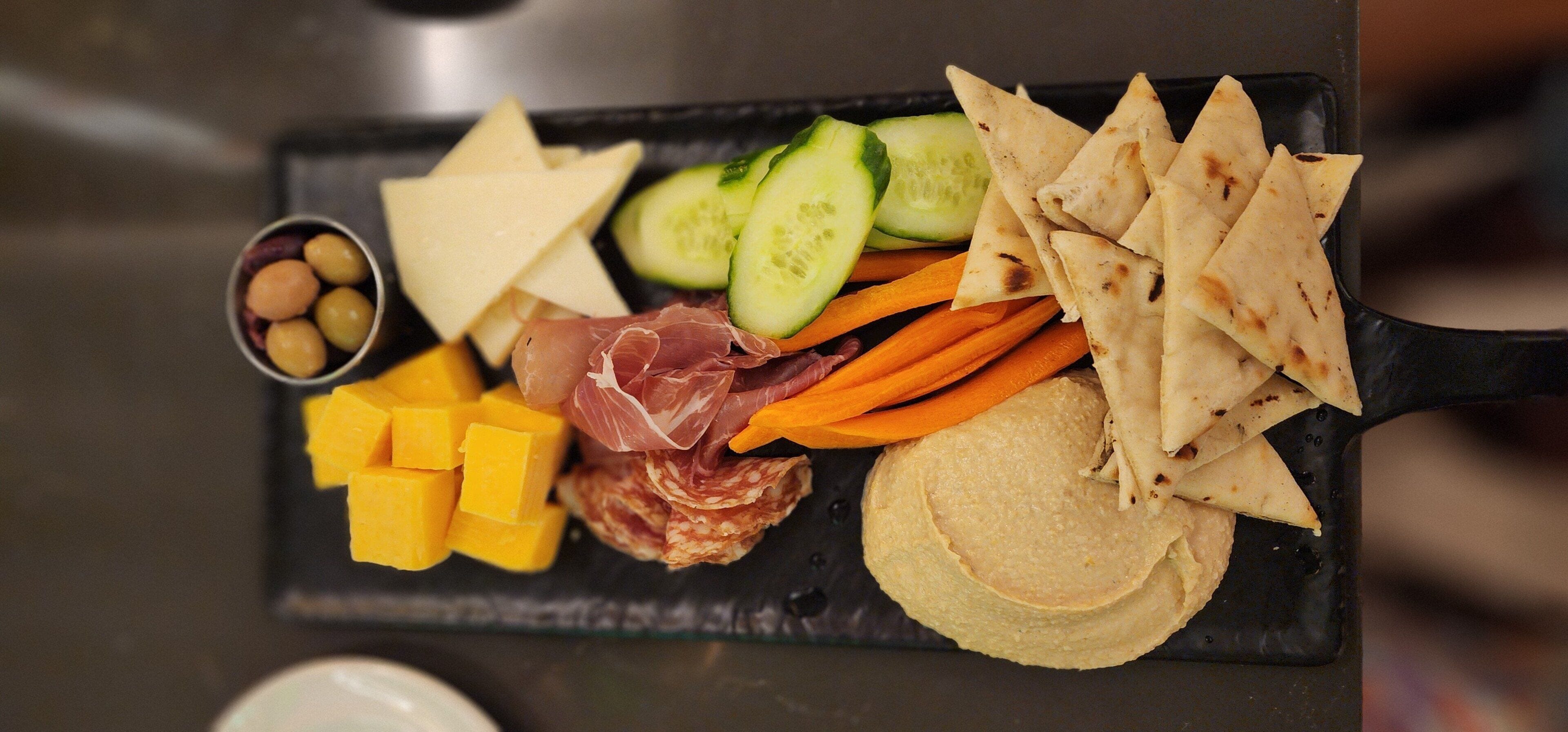 A black tray with various food items including cheese, prosciutto, cucumbers, carrots, and flatbread.
