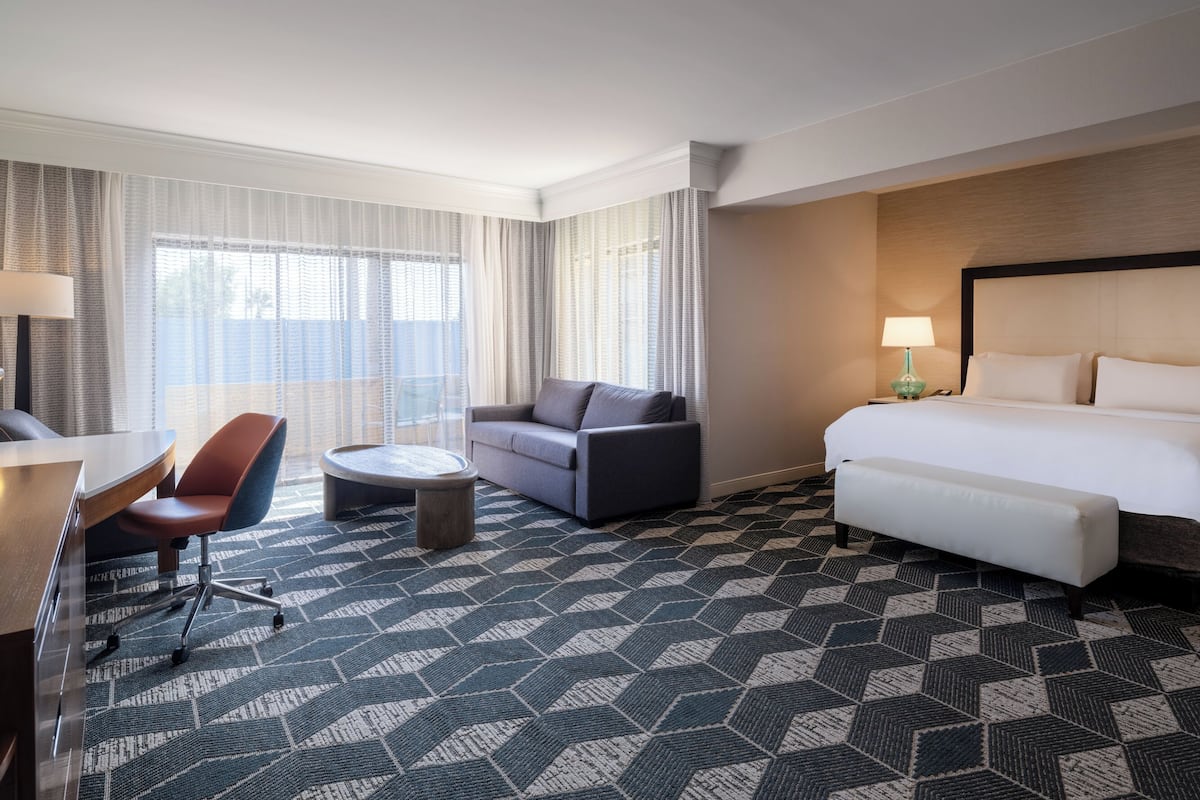 Una habitación de hotel con cama, sofá, sillón, una mesita y una lámpara.