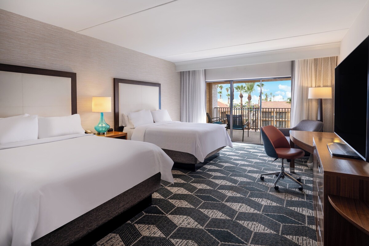 Habitación de hotel con dos camas, un escritorio, un televisor y un balcón con vistas a palmeras.