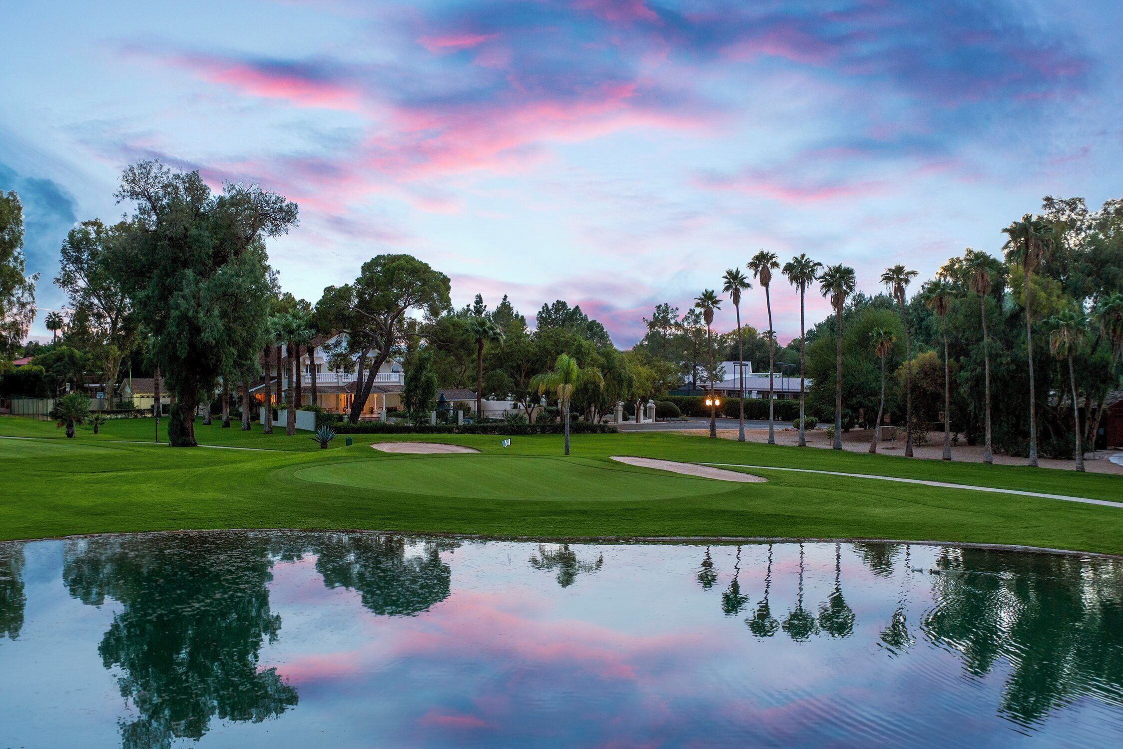 Un campo de golf con un estanque que refleja el cielo, rodeado de árboles y edificios.
