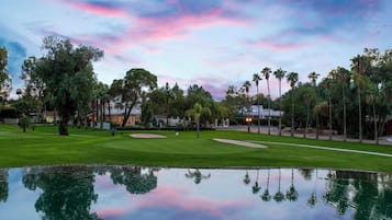 Un campo de golf con un estanque que refleja el cielo, rodeado de árboles y edificios.