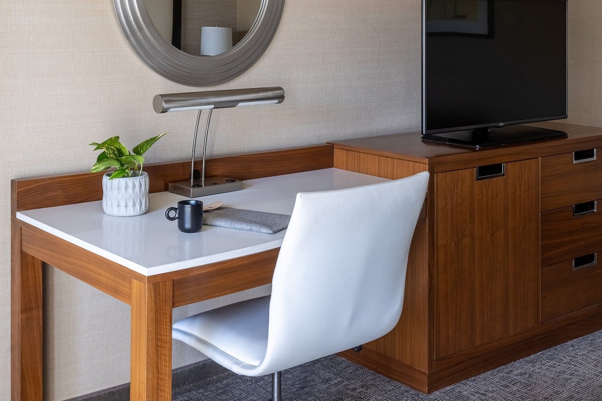 Habitación de hotel con un escritorio de madera, una silla blanca, un televisor, un espejo y una planta en maceta.