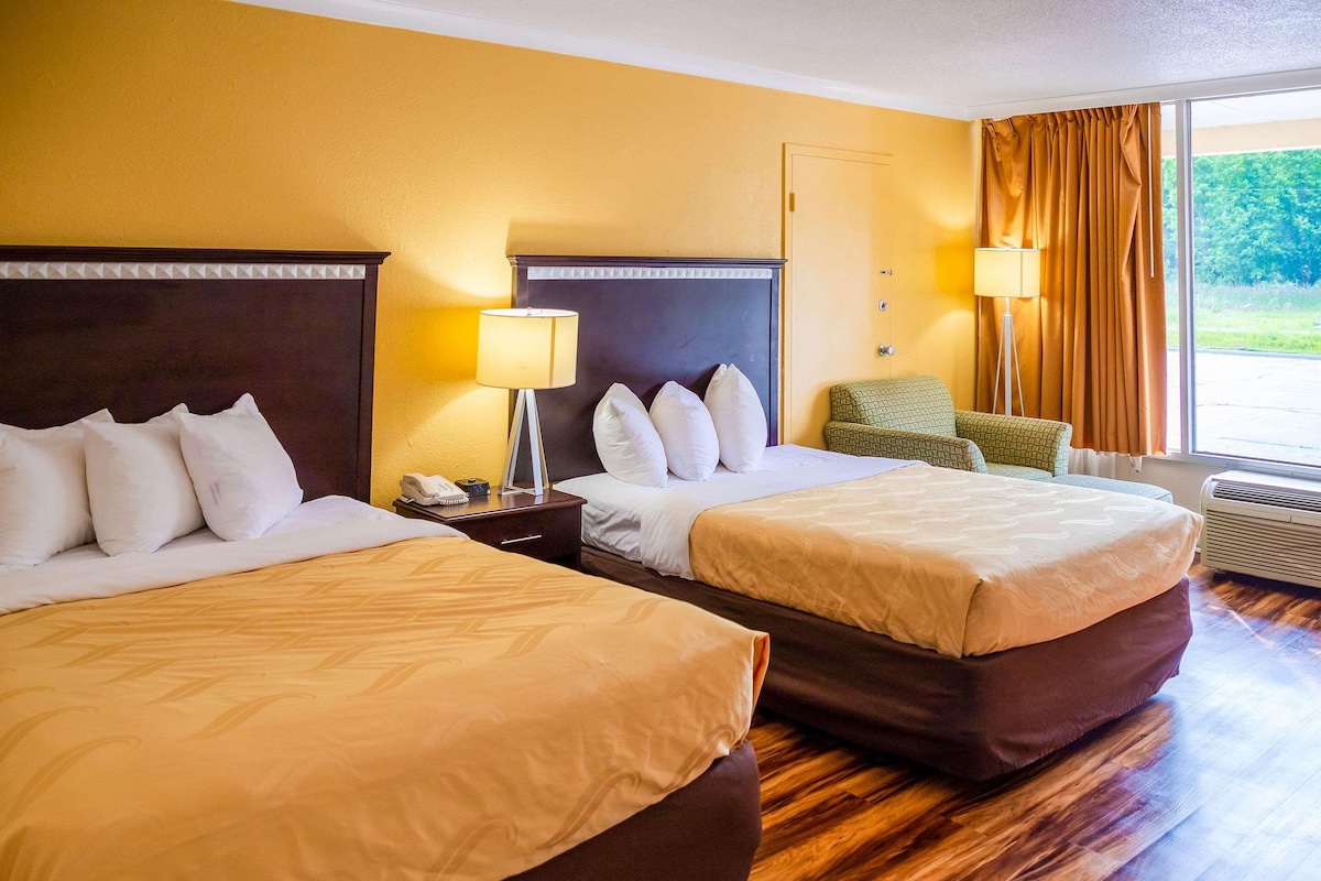 A hotel room with two beds, a nightstand with a lamp, a chair, and a view of the outdoors.