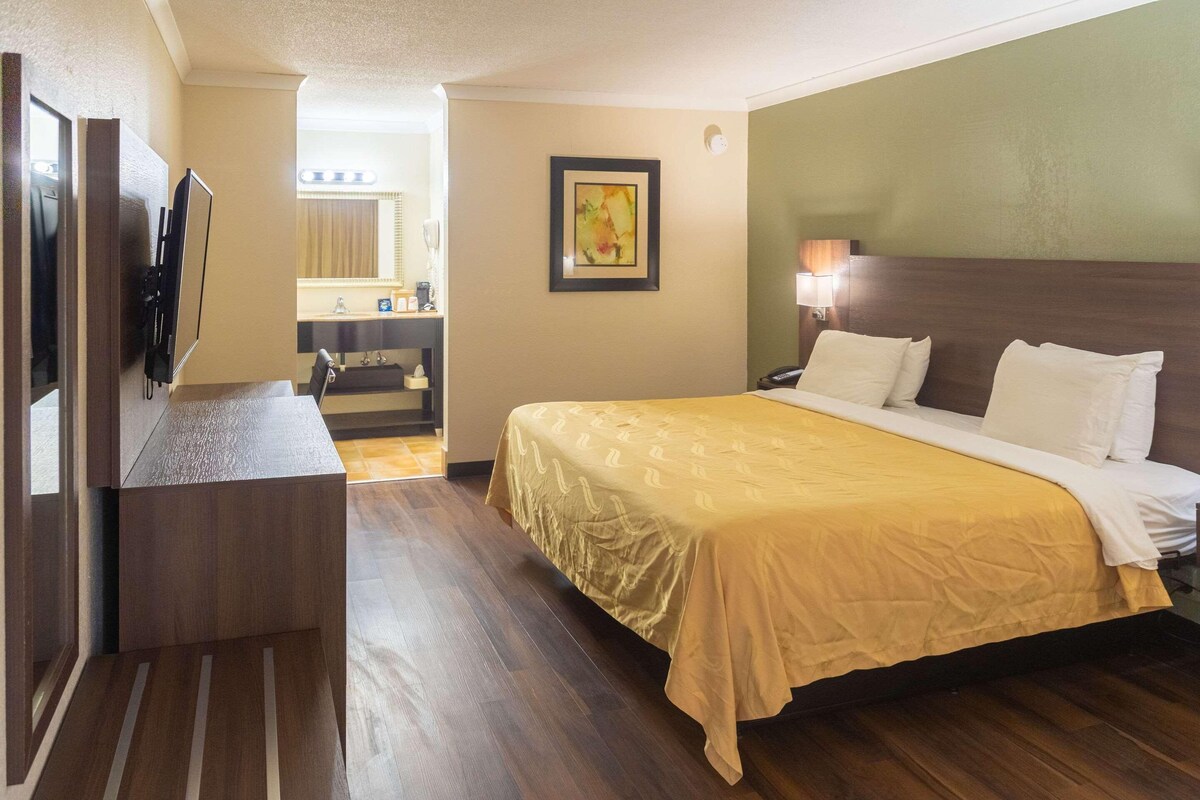 A hotel room with a large bed, a desk with a flat-screen TV, a bathroom with a mirror and sink, and a framed picture on the wall.