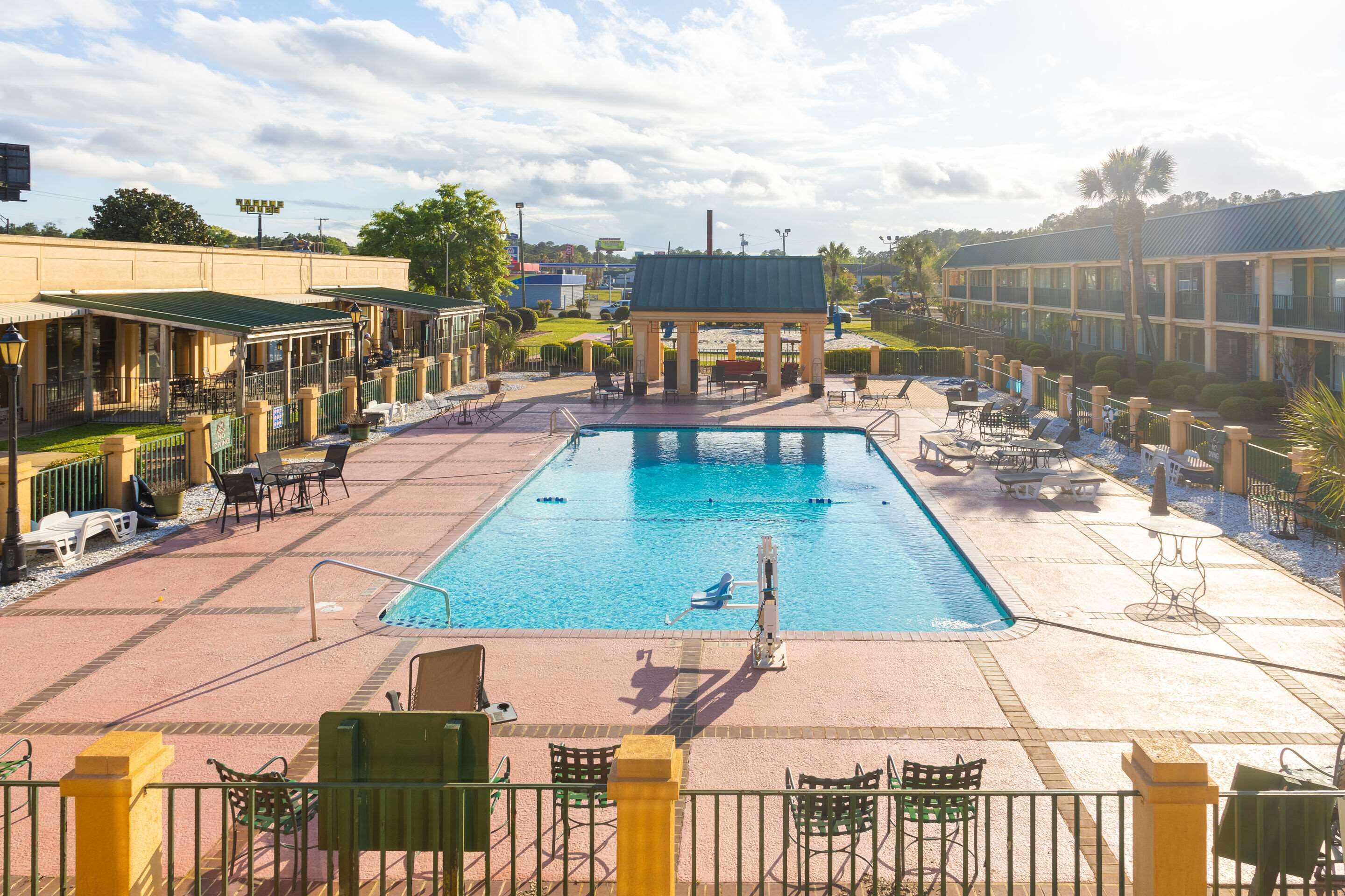 A swimming pool with lounge chairs and umbrellas, surrounded by a fence and a building with a green roof.