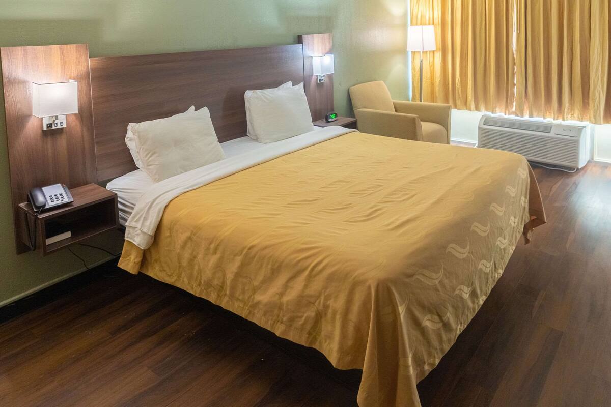A hotel room with a large bed, bedside lamps, a chair, and an air conditioning unit.