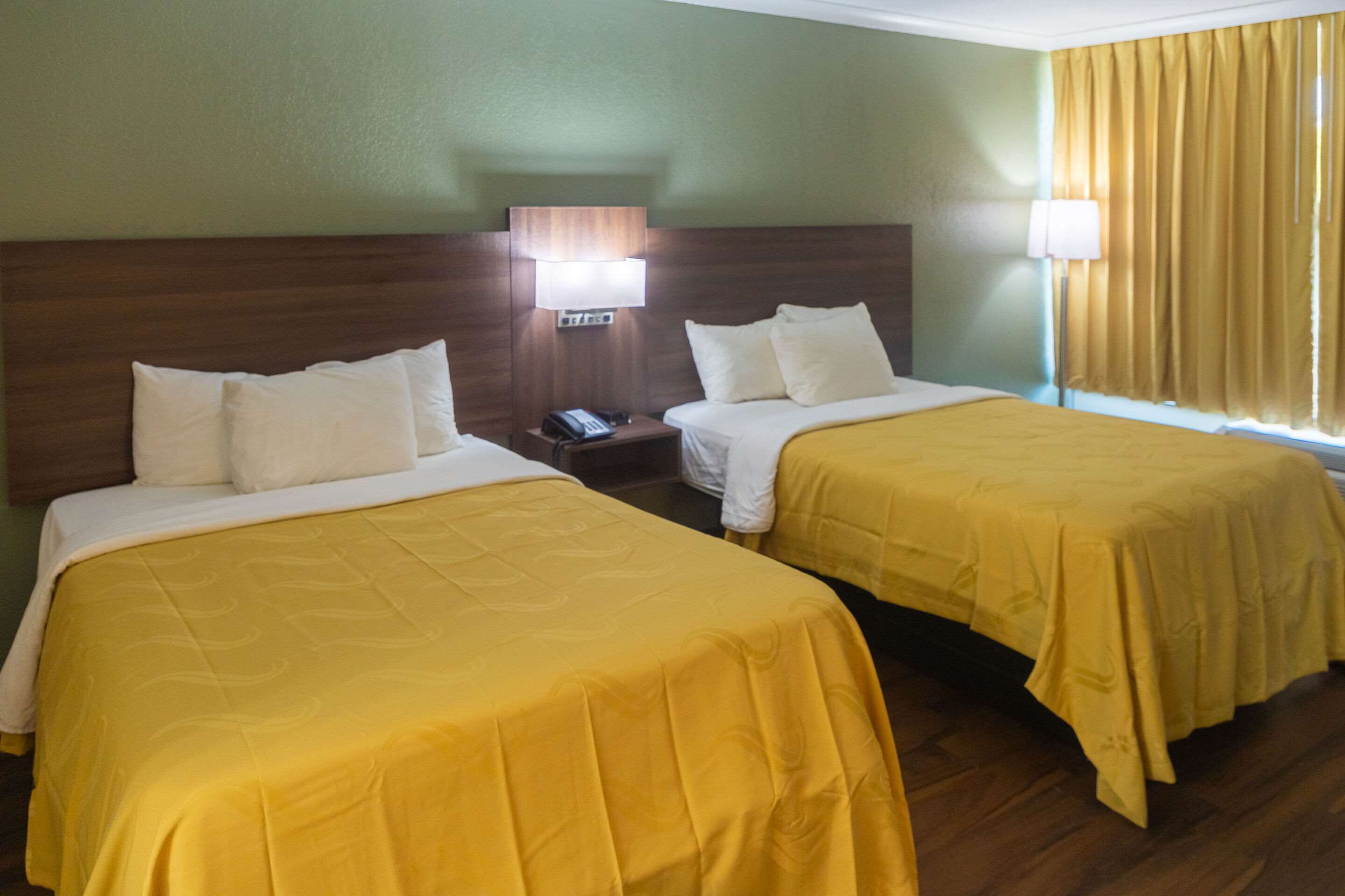 Habitación de hotel con dos camas, cada una con cubrecama amarillo y almohadas blancas. Se observa un mesita de noche con una lámpara y un teléfono. La habitación tiene un cabecero de madera y una ventana con cortinas amarillas.