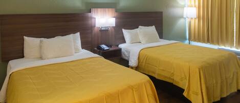 Habitación de hotel con dos camas, cada una con cubrecama amarillo y almohadas blancas. Se observa un mesita de noche con una lámpara y un teléfono. La habitación tiene un cabecero de madera y una ventana con cortinas amarillas.