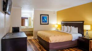 Une chambre d’hôtel avec un lit, des lampes de chevet, un téléviseur posé sur un meuble, un tableau accroché au mur et une salle de bain visible à travers une porte ouverte.