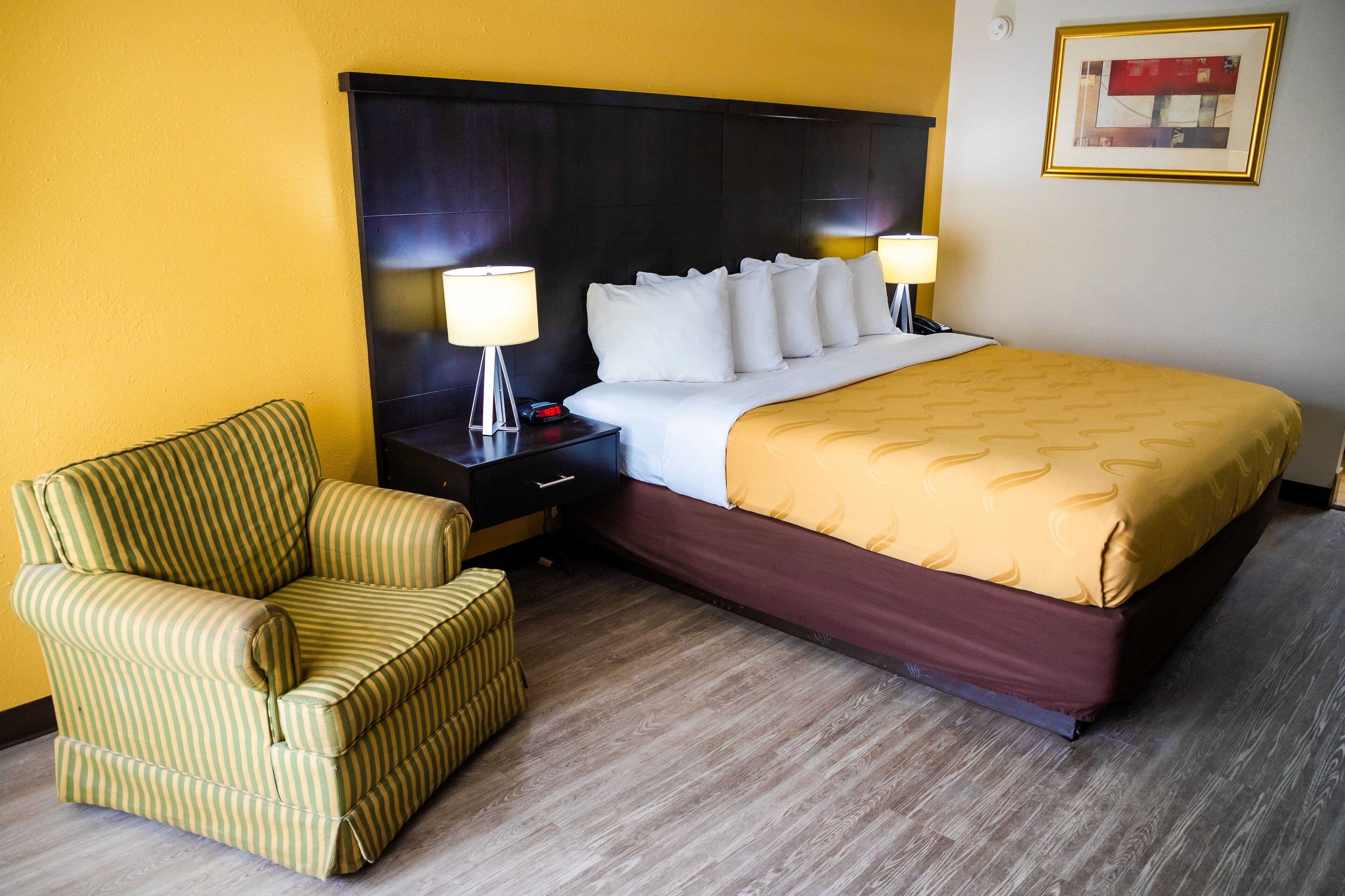 Una habitación de hotel con una cama grande, una mesita de noche con lámpara, un sillón a rayas y un cuadro en la pared.
