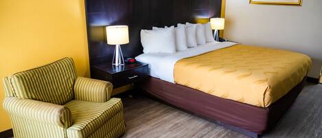 Una habitación de hotel con una cama grande, una mesita de noche con lámpara, un sillón a rayas y un cuadro en la pared.