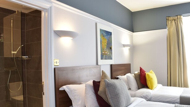 A hotel room with a shower, two beds with multiple pillows, a painting on the wall, and a window with curtains. - Royal Oxford Hotel (Oxford)