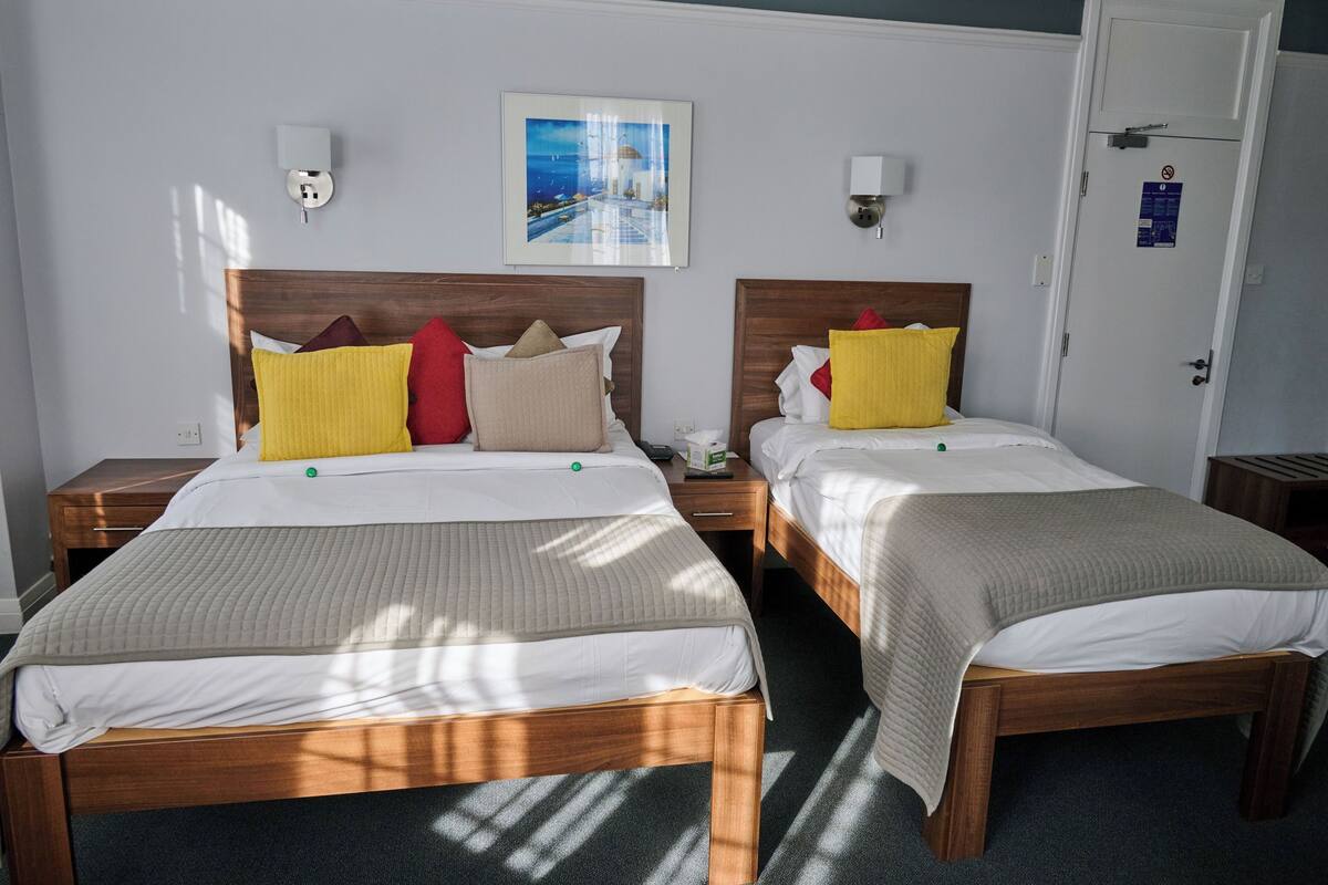 Une chambre d’hôtel avec deux lits, chacun doté d’une tête de lit en bois, de draps blancs et de plusieurs oreillers. Un tableau encadré est accroché au mur au-dessus des lits, et une porte porte une pancarte.