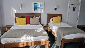 Een hotelkamer met twee bedden, elk met een houten hoofdbord, wit beddengoed en diverse kussens. Boven de bedden hangt een ingelijst schilderij en er is een deur met een bordje op.