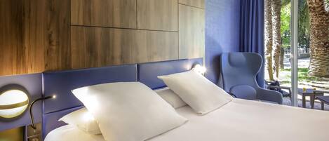 Ein Hotelzimmer mit einem großen Bett, einer Sitzecke mit einem Sessel, einem Holz-Kopfteil und Blick auf Palmen.