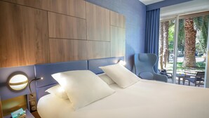 Ein Hotelzimmer mit einem großen Bett, einem Nachttisch mit Lampe, einem Sessel und Blick auf Palmen durch das Fenster.
