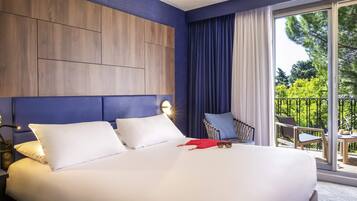 A hotel room with a large bed, a headboard with built-in lighting, a view of the outdoors through a sliding glass door, and a seating area with a chair and table.
