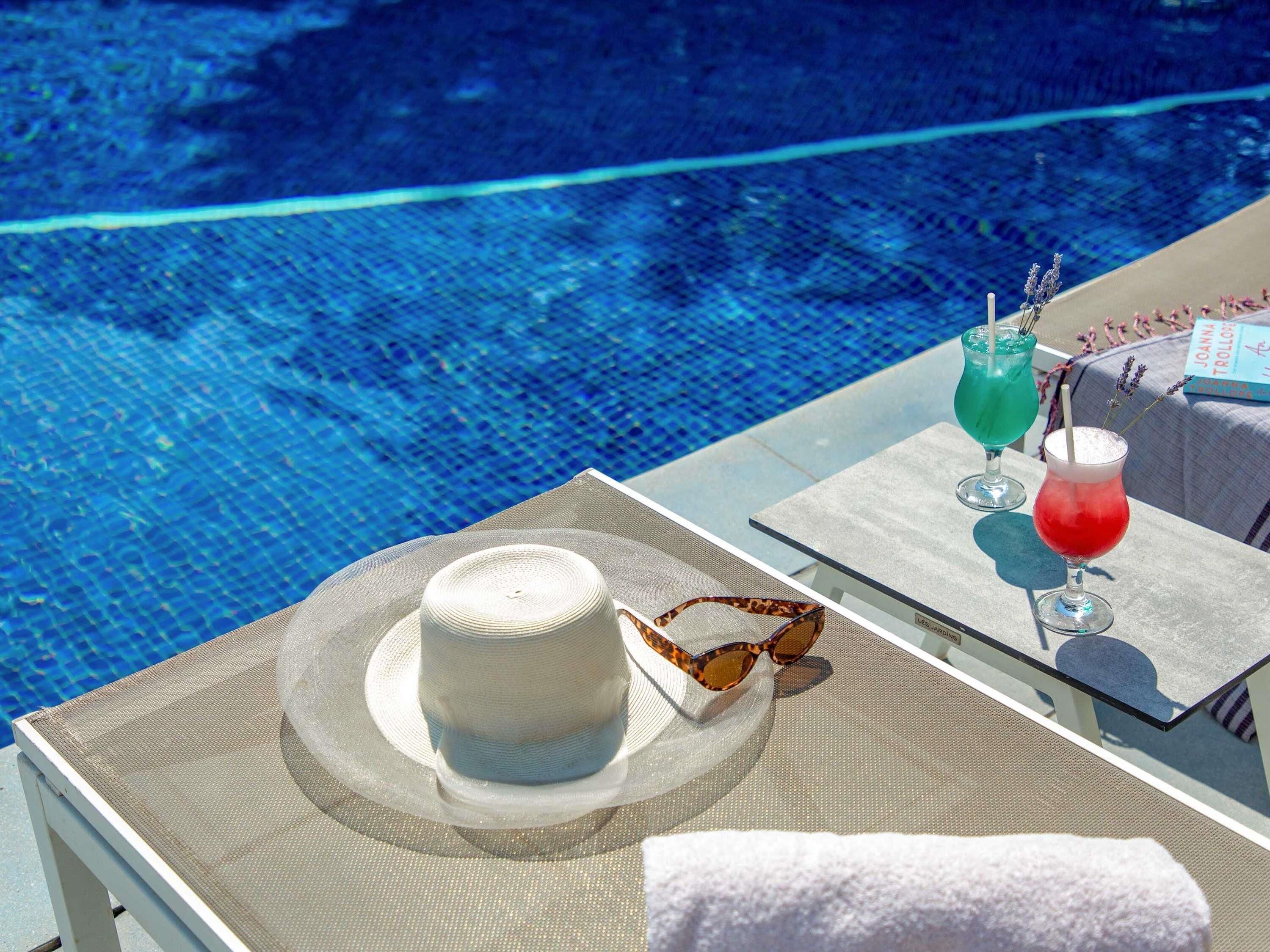 Escena junto a la piscina con un sombrero, gafas de sol y dos bebidas sobre una mesa.