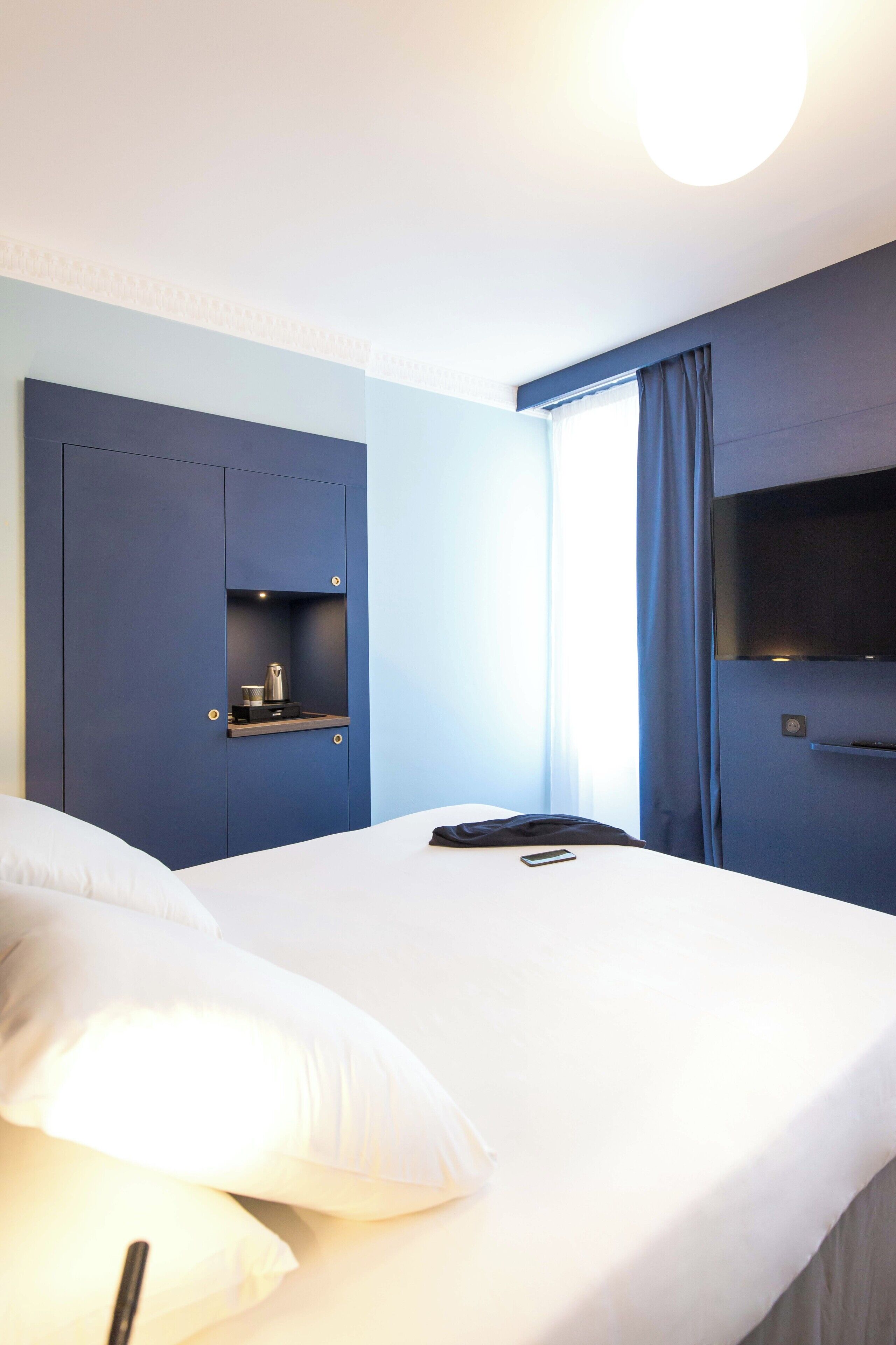 Une chambre d’hôtel avec un lit, une petite étagère avec une bouilloire, une télévision et une fenêtre avec des rideaux.