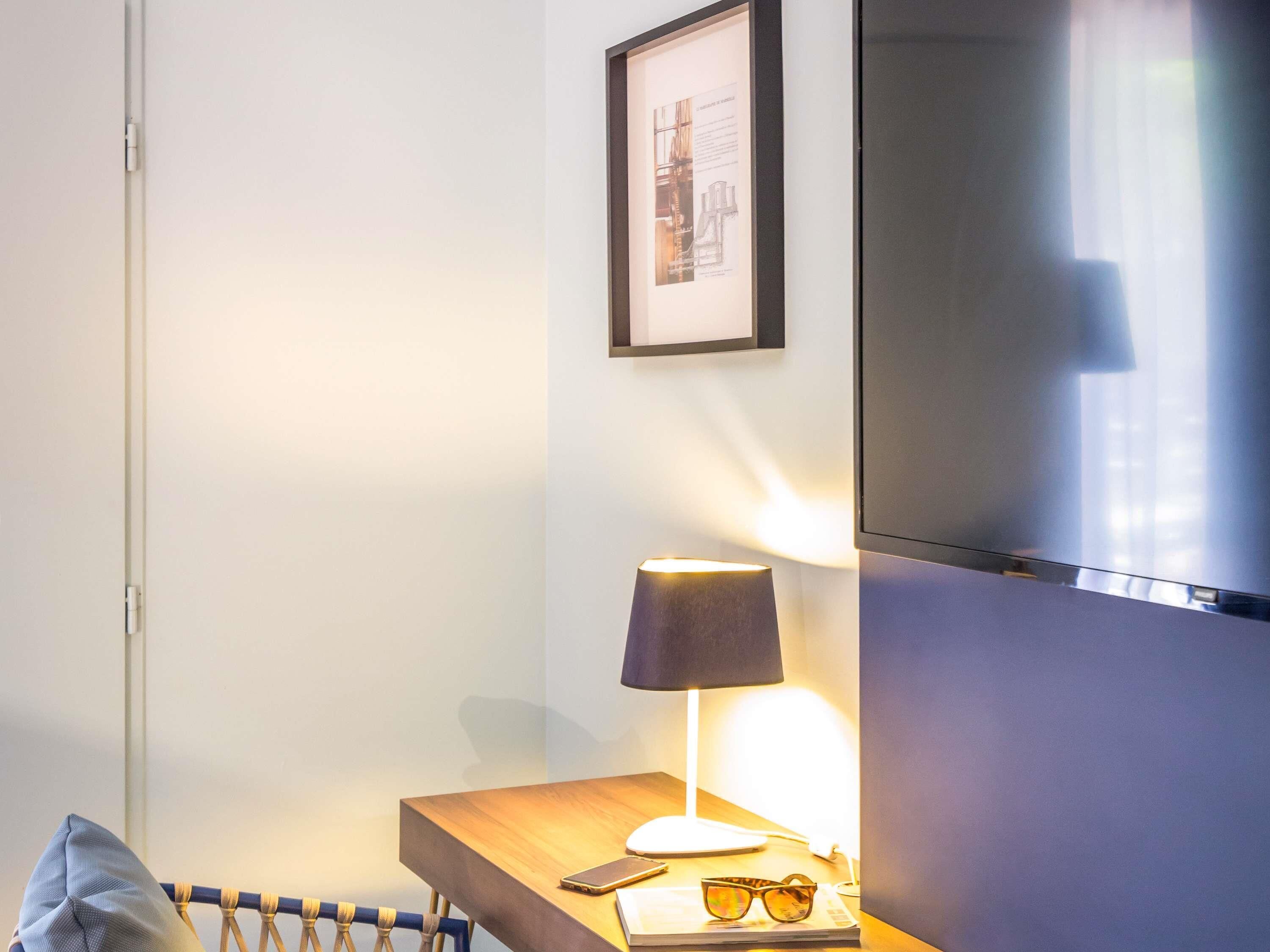 Habitación de hotel con cama, mesita de noche con lámpara, televisor y un cuadro en la pared.