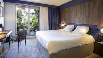 A hotel room with a bed, a desk with a laptop, a chair, and a balcony with a view of greenery.