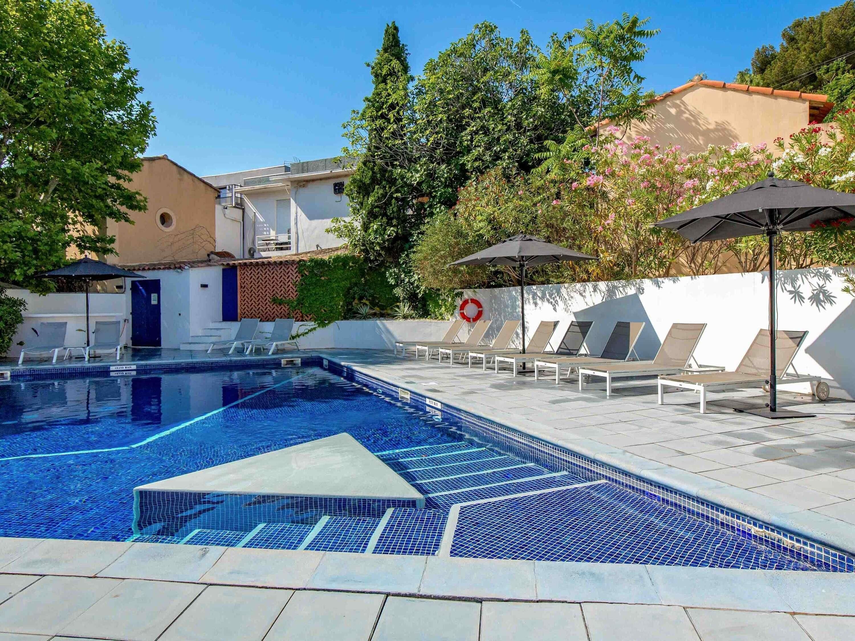 A swimming pool with lounge chairs and umbrellas, surrounded by buildings and trees.