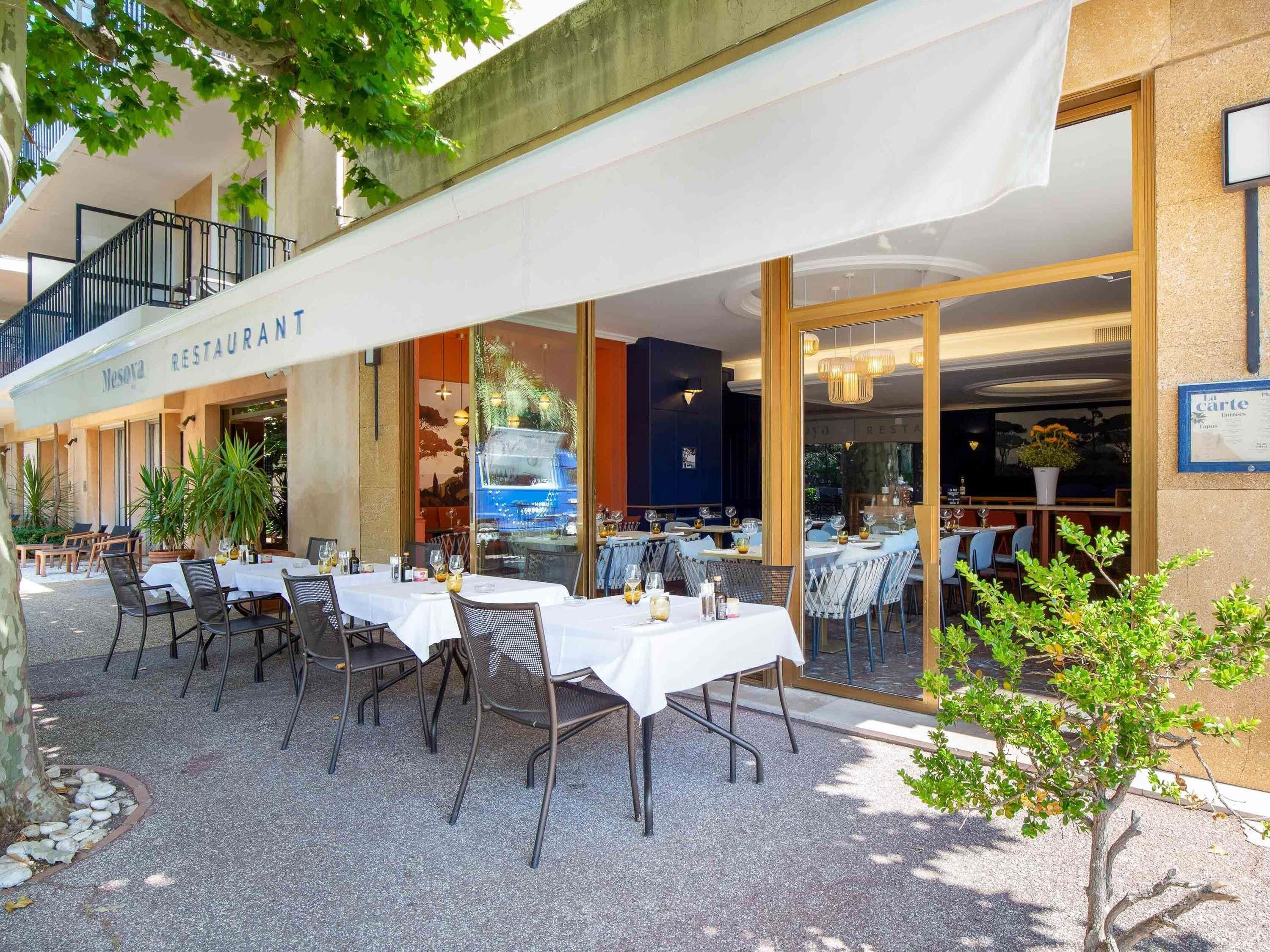 Un área de comedor al aire libre con mesas y sillas, un paseo techado y la entrada de un edificio.