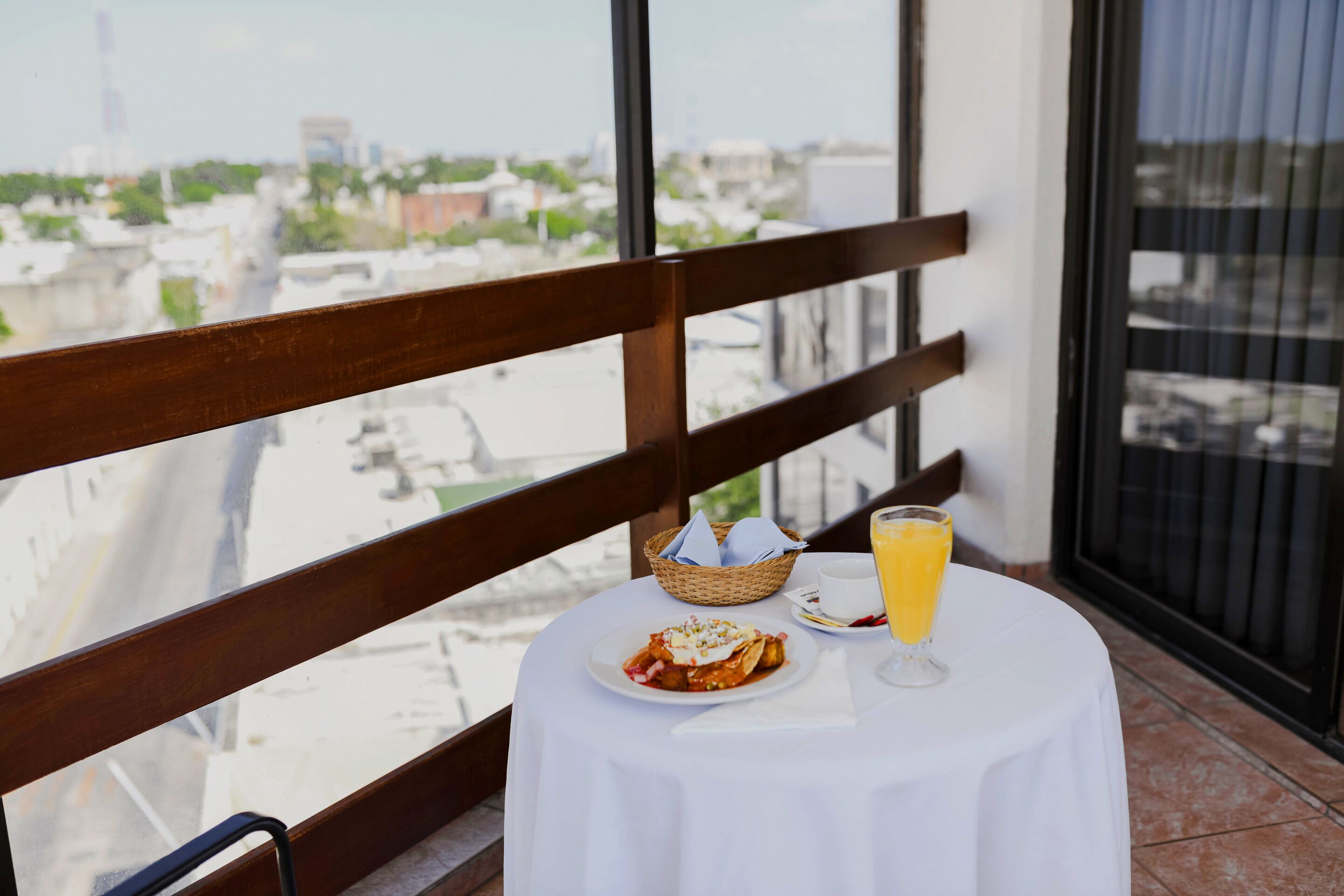 A balcony with a table set for a meal, a wicker basket, a glass of orange juice, and a view of the cityscape.