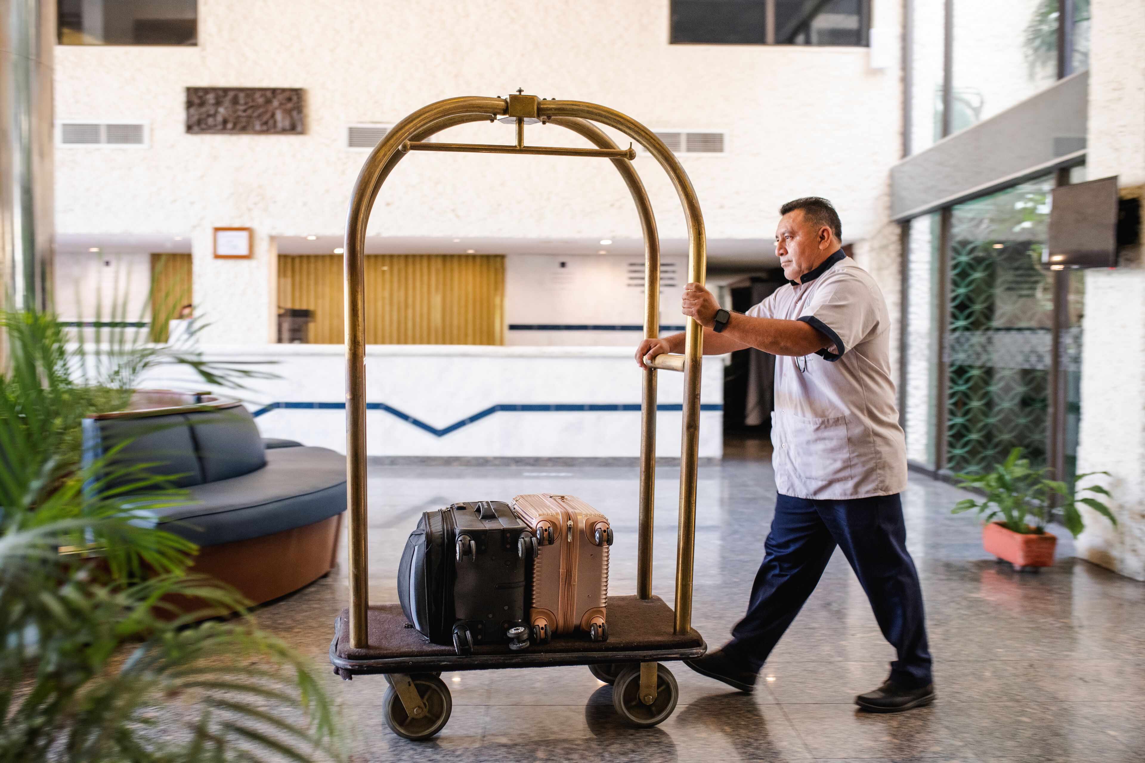 Un homme pousse un chariot à bagages rempli de valises dans un hall d’accueil où se trouvent un comptoir de réception, des sièges et des plantes en pot.