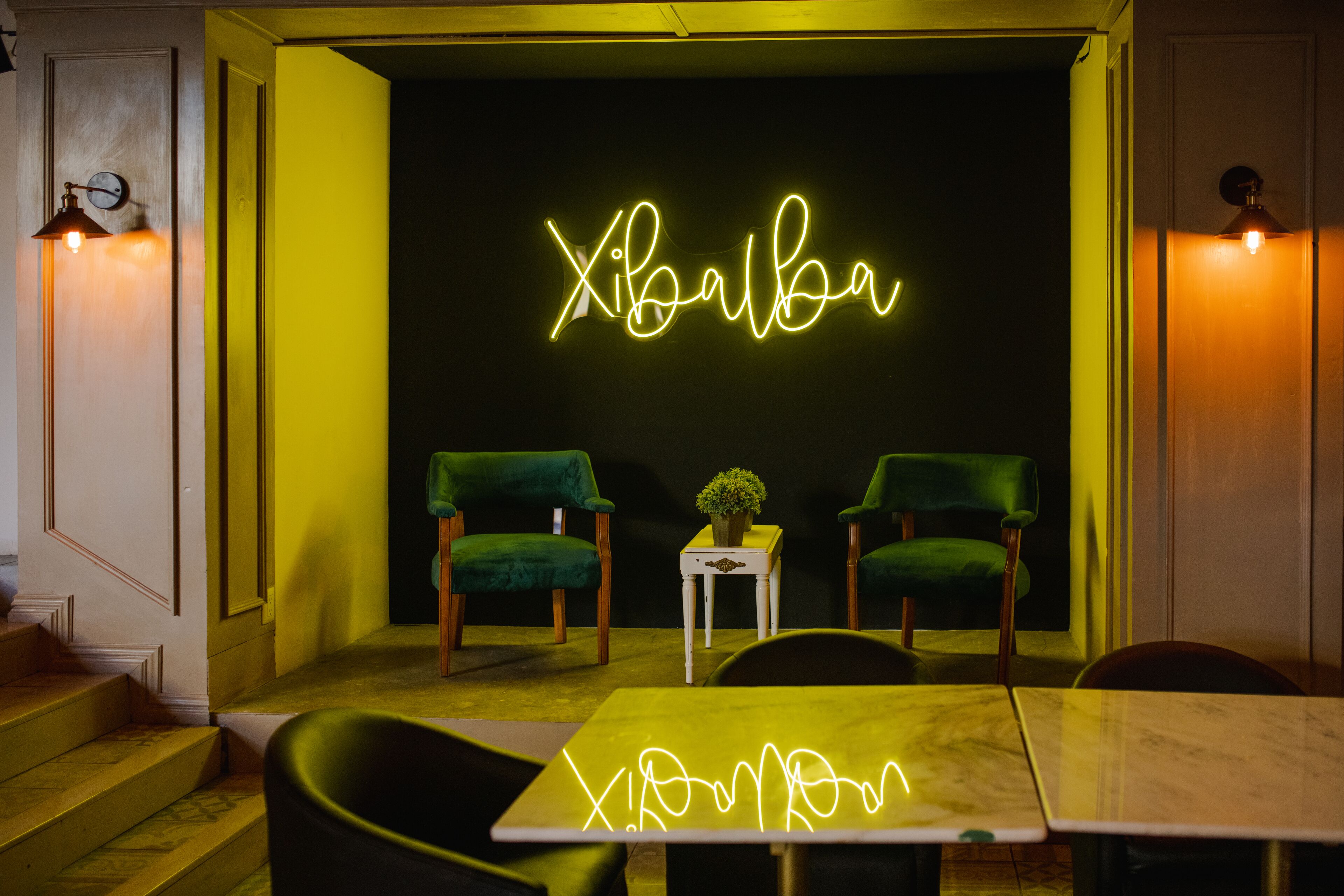 A room with a neon sign reading "Xibalba," two green velvet chairs, a small table with a plant, and a marble table in the foreground.