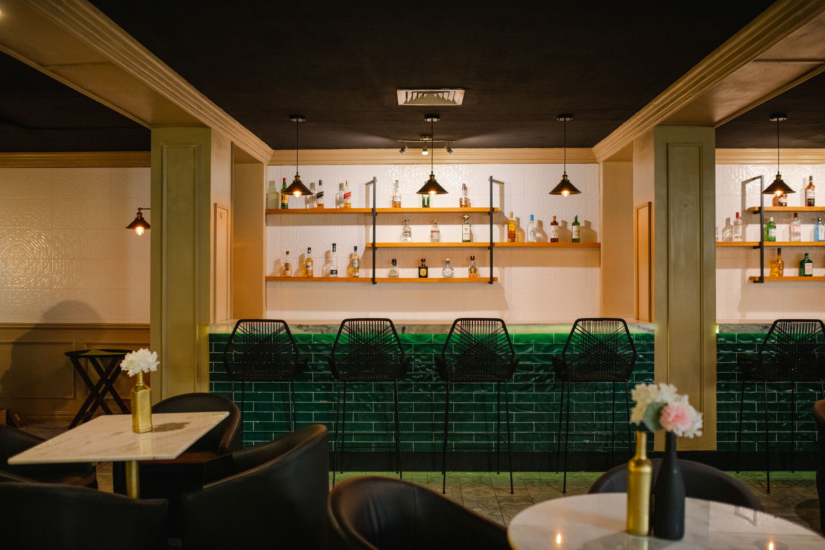 A bar area with shelves of bottles, black chairs, and a green tiled wall.