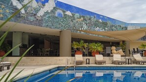Uma área de piscina com espreguiçadeiras e plantas em vasos, um mural com animais selvagens e uma piscina azul cristalina.