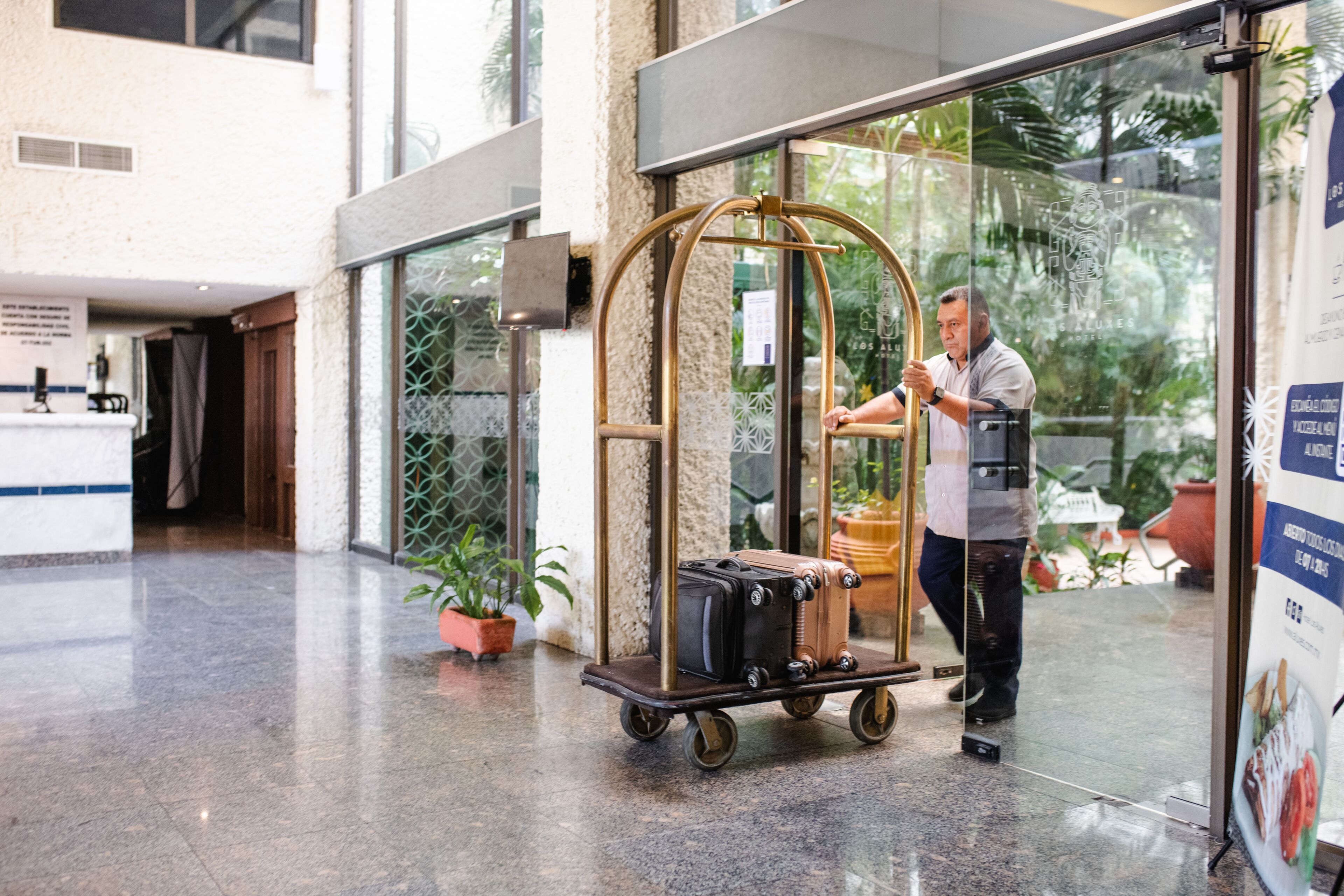 A luggage cart with suitcases in a hotel lobby with a glass door and potted plants.
