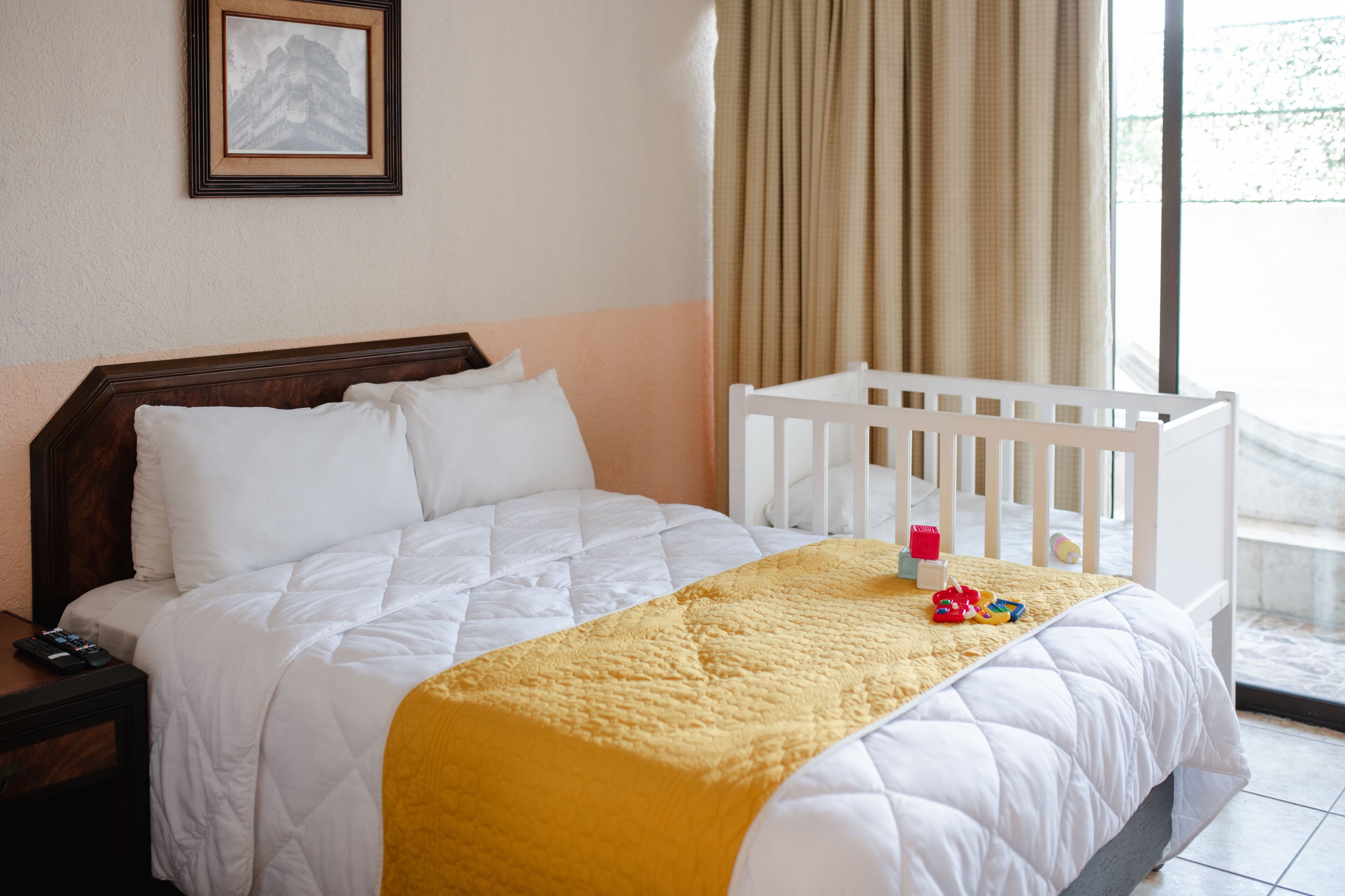 Ein Hotelzimmer mit einem Bett, einem weißen Babybett, einer gelben Decke und einem gerahmten Bild an der Wand.