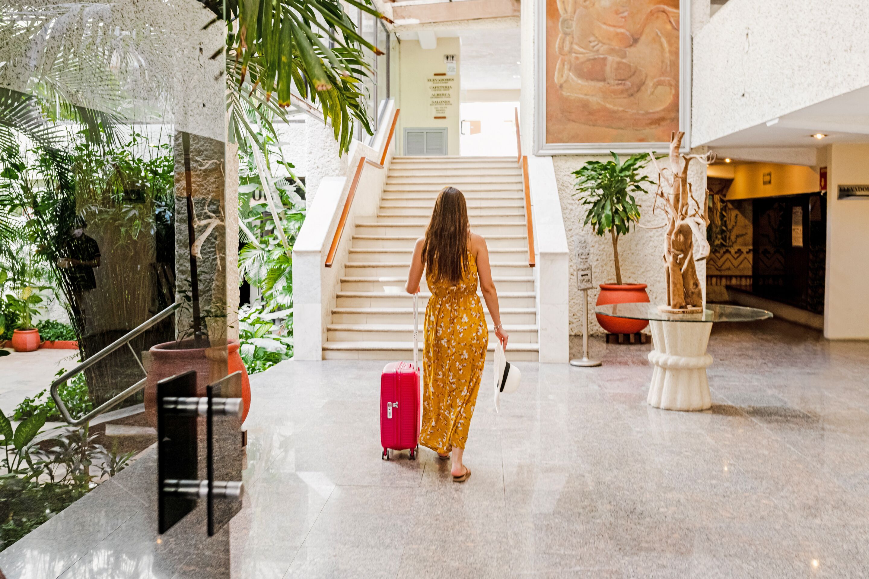 A woman in a yellow floral dress walking with a red suitcase in a lobby with a staircase, potted plants, and a large wall mural.