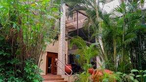 Um pátio exuberante com uma escadaria que leva à entrada de um edifício, rodeado por vegetação tropical densa e grandes vasos de terracota.