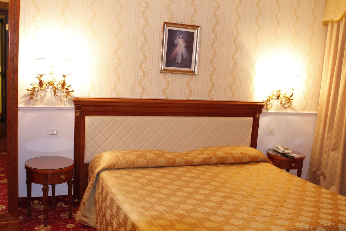 Habitación de hotel con cama, dos mesitas de noche, lámpara y un cuadro en la pared.