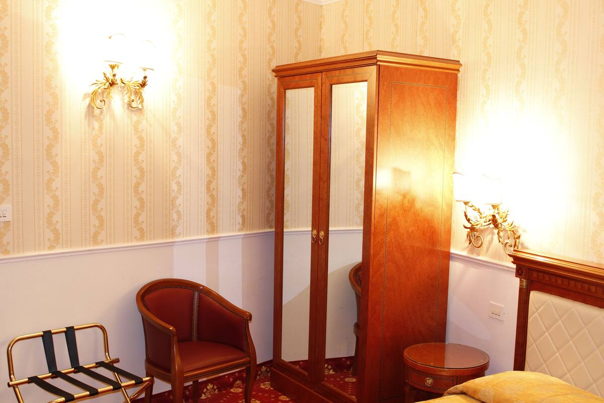 Une pièce comprenant une armoire en bois, une chaise rouge, une petite table ronde et un lit avec une tête de lit matelassée.