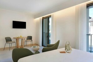 A modern hotel room with a flat-screen TV mounted on the wall, a dining area with a round table and chairs, and a large window with curtains. - Hotel Lloyd Milan (Milan)