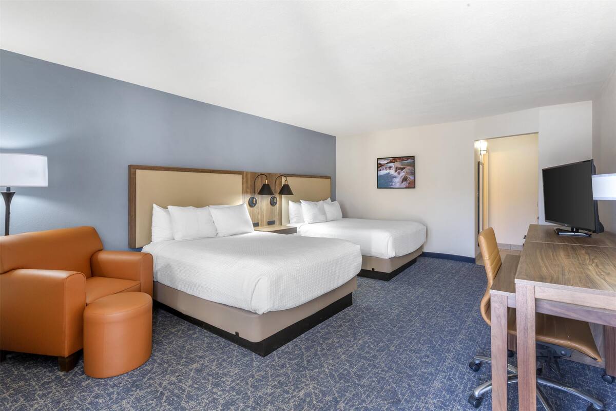 A hotel room with a double bed, two bedside tables, a desk with a chair, an orange armchair, and a TV.