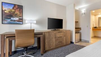 A hotel room with a wooden desk, a television, a chair, a bed, a bathroom with a mirror, and a painting of a canyon.
