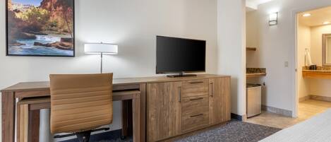A hotel room with a wooden desk, a television, a chair, a bed, a bathroom with a mirror, and a painting of a canyon.