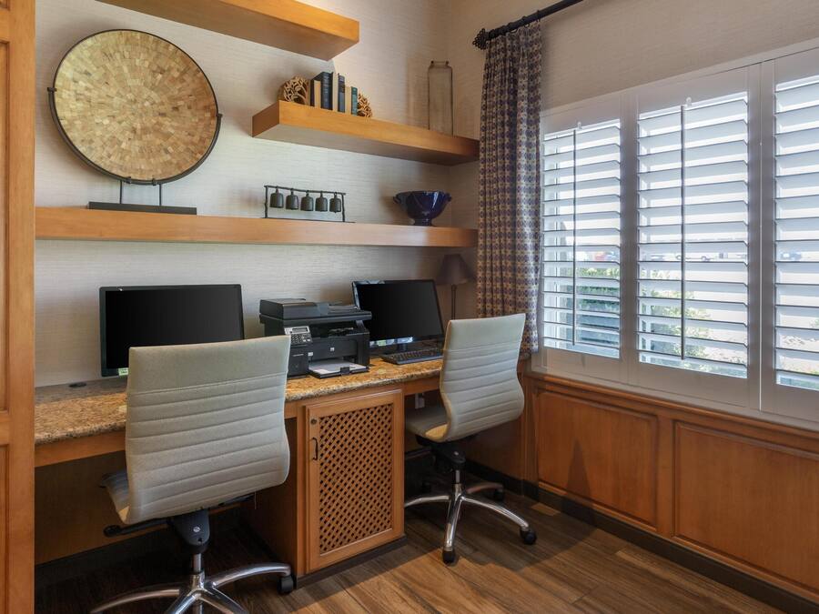 Un bureau à domicile équipé de deux bureaux, d’ordinateurs et de chaises de bureau. On y trouve des étagères ornées d’objets décoratifs et une fenêtre avec des stores.