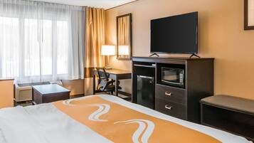 Une chambre d’hôtel comprenant un lit, un bureau avec une chaise, une télévision, une armoire et une fenêtre avec des rideaux.