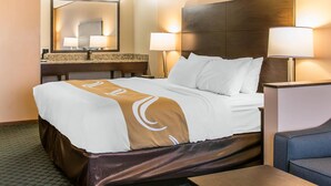 A hotel room with a large bed, a nightstand, a lamp, a mirror, and a bathroom counter. - Quality Inn & Suites Houghton Downtown (Houghton)