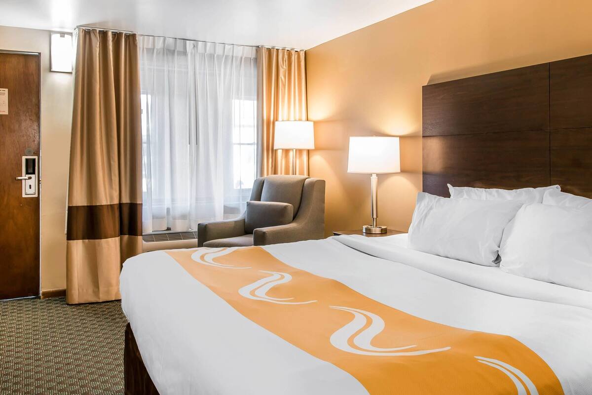 A hotel room with a large bed, a chair, a lamp, and a window with curtains.
